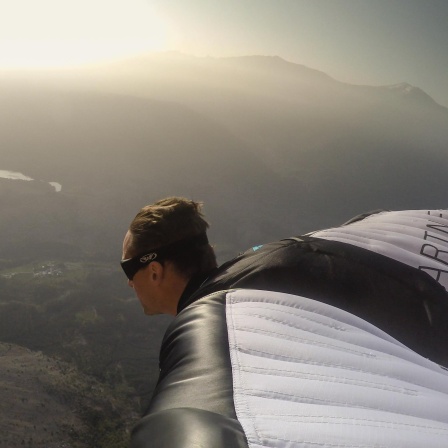 Der Basejumper Maximilian Werndl beim Flug in einem drachenartigen Anzug (Wingsuit) vor einem Bergpanorama im Gegenlicht.