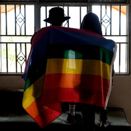 Ein Paar sitzt mit dem Rücken zur Kamera in einem Gebäude vor einem Fenster. Beide haben sich eine Pride-Flagge um die Schultern gelegt.