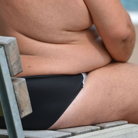 Ein übergewichtiger Mensch sitzt in einem Freibad auf einer Bank