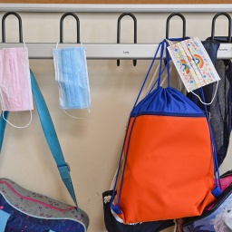 Masken hängen zusammen mit Taschen und Rucksäcken an Kleiderhaken in einem Klassenraum einer Grundschule.
