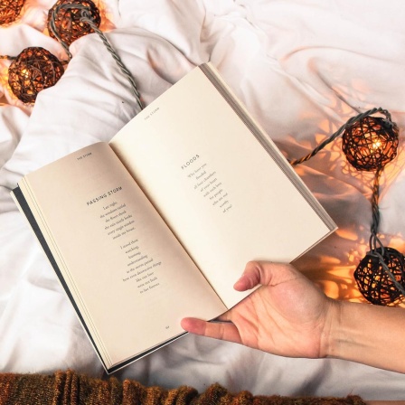 Draufsicht auf ein aufgeschlagenes Buch. Die Beine der Leserin sind sichtbar und in herbstliche, lange Strümpfe gekleidet. Außerdem sieht man eine Tasse Kaffee und Leuchtkugeln. Die Atmosphäre ist herbstlich und behaglich.