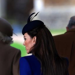 Prinzessin Kate ist schräg von hinten zu sehen. Sie trägt einen blauen Hut mit Federn. Vor ihr die dunklen Silhouetten von König Charles und Königin Camilla.