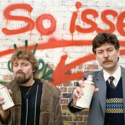 Jürgen von der Lippe und Gerd Dudenhöfer stehen mit Spraydosen in der Hand vor einer Hauswand, auf der in roter Farbe der Schriftzug "So isses" prangt (1984)