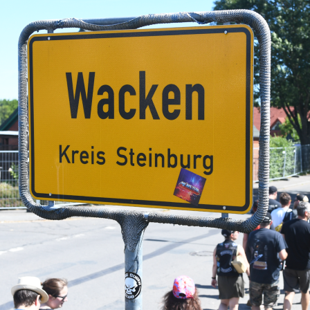Wacken Besucher laufen an dem Dorfschild "Wacken Kreis Steinburg" durch die Straßen