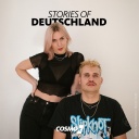 Ein Mann sitzt mit schwarzem T-Shirt auf einem Stuhl vor weißem Hintergrund, am Stuhl lehnt eine Frau, beide schauen in die Kamera.