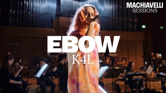 Ebow steht vor dem WDR-Funkhausorchester mit einem Mikrofon in der Hand, über dem Bild liegt der Schriftzug "Ebow K4L"