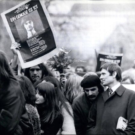 Trauerzug mit Menschen aus dem alternativen Milieu in typischer Kleidung und Anmutung. Einer von ihnen hält ein Schild mit dem Bild von Holger Meins hoch, auf dem steht "Ein Genosse ist tot".