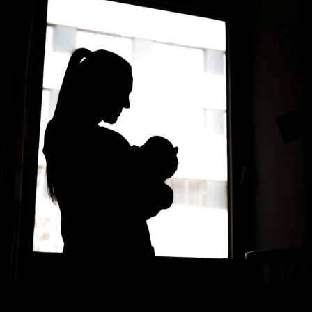 Eine Frau steht mit einem Neugeborenen am Fenster der Klinik.