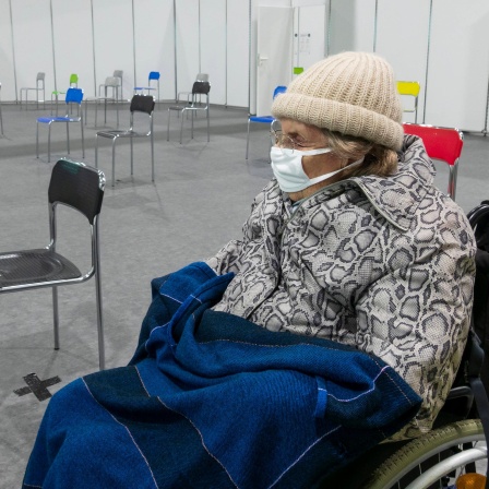Eine ältere Fraue mit Schutzmaske und warmer Kleidung sitzt auf einem Rollstuhl in einem Impfzentrum in Spanien, 2021.