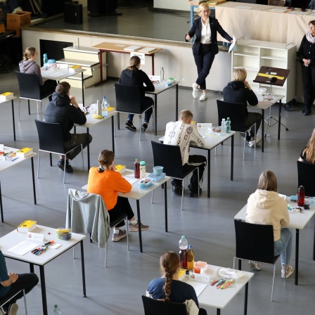 In der Aula in einem Gymnasium beginnt die schriftliche Abiturprüfung in Deutsch - Schülerinnen und Schüler sitzen an Einzeltischen und werden beaufsichtigt.
