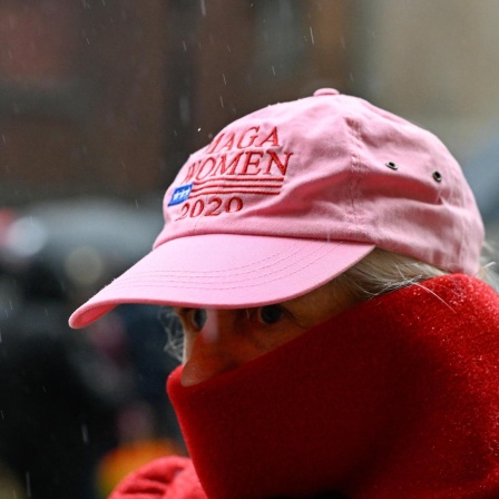 Eine Frau trägt eine pinkte Baseballmütze mit der Aufschrift "Maga Women" und einen Schal vor dem Gesicht, um sich vor der Kälte zu schützen.