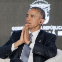 Barack Obama faltet die Hände vor dem Gesicht zusammen, während er auf einer Konferenz einen Kopfhörer im Ohr hat.