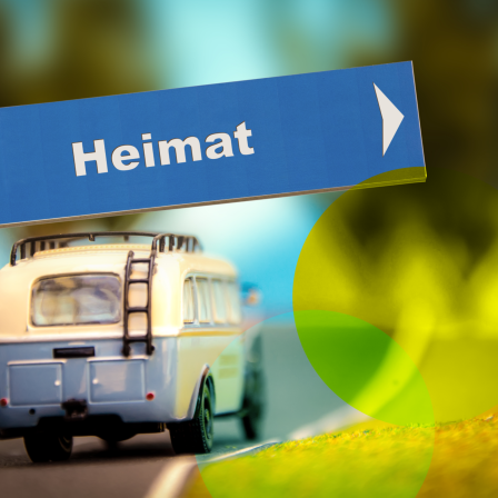 Das Bild zeigt ein Verkehrsschild, das mit dem Wort „Heimat“ beschriftet ist. Das Schild ist blau und hat einen weißen Pfeil, der nach rechts zeigt. Es steht an einem Straßenrand, und im Hintergrund sieht man ein Modell eines nostalgischen, hellblauen Reisebusses mit einem Gepäckträger auf dem Dach, der eine kurvige Straße entlangfährt.