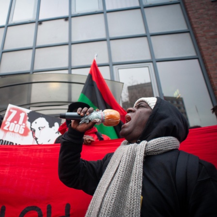 Mouctar Bah steht mit einem Mikrofon in der Hand vor einem roten Banner. Dahinter weiter Aktivisten, die Oury Jalloh gedenken.