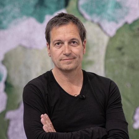 Porträt von Dieter Nuhr mit verschränkten Arnmen vor einem Kunstwerk 