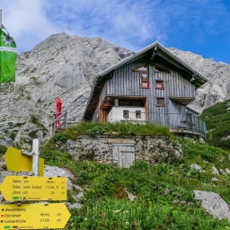 Eine Bergsteigerhütte in den Alpen, davor gelbe Schilder und eine grüne Flagge, dahinter die Alpen