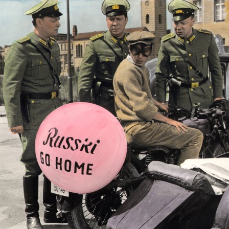 Filmszene aus Billy Wilders "Eins, Zwei,Drei", an einem Motorrad bläst sich ein Luftballon mit der Aufschrift: Russki Go Home auf, das am Grenzübergang in Berlin kontrolliert wird