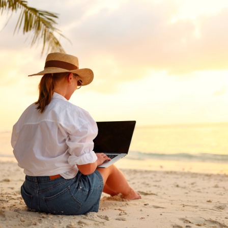 Junge Frau mit Strohhut arbeitet am Strand mit Notebook.
