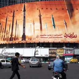 Ein Mann überquert eine Straße, während Autofahrer an einer Werbetafel vorbeifahren, auf der iranische ballistische Raketen abgebildet sind.