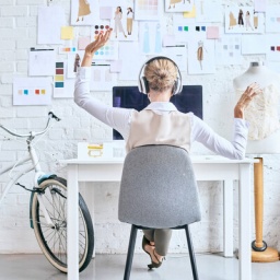 Eine Frau sitzt an einem Schreibtisch, vor ihr an der Wand hängen viele Zettel, ein Moodboard, neben dem Schreibtisch ein Fahrrad und Stoffrollen.