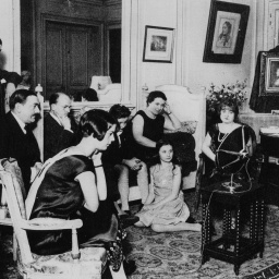 Eine 8-köpfige Familie sitzt im Wohnzimmer um eine Radiogerät, das im Zentrum des Raumes steht.