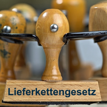 Ein Holzstempel mit der Aufschrift "Lieferkettengesetz" sowie weitere Stempel in einem Stempelkarussell stehen auf einem Schreibtisch.