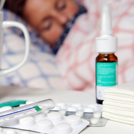 Medikamente und ein Fieberthermometer liegen auf einem Nachttisch - im Hintergrund liegt eine Frau mit Erkältung im Bett und schläft.