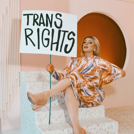 Die Dragqueen und Politikerin Maebe A. Girl mit einem Schild auf dem "Trans Rights" steht vor einer orangefarbenen Kulisse.
