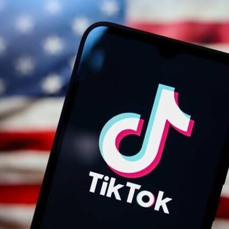 Tiktok-Logo auf einem Handydisplay, im Hintergrund die US-amerikanische Flagge