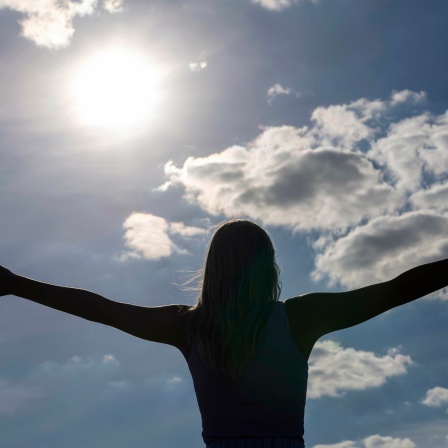 Das Foto zeigt eine Frau mit ausgestreckten Armen, vor ihr ist der sonnige Himmel zu sehen
