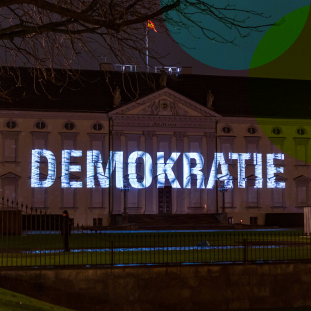 Das Bild zeigt eine nächtliche Projektion des Wortes "DEMOKRATIE" auf die Fassade eines historischen Gebäudes, das Schloss Bellevue in Berlin, den Amtssitz des deutschen Bundespräsidenten.