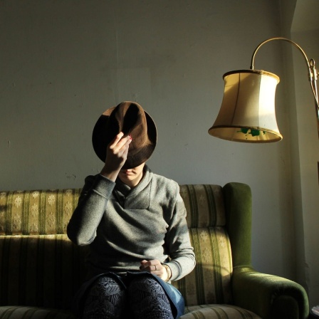 Eine Person sitzt auf einem altmodischen Sofa neben einer Stehlampe. Das Gesicht verdeckt sie mit einem Hut. 