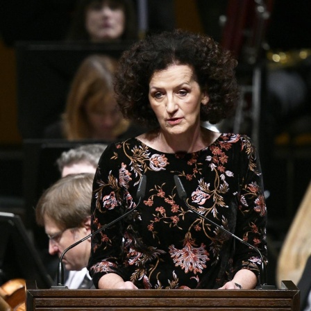 Maja Haderlap spricht auf einem Podium. Sie hat dunkelbraune Locken und trägt ein schwarzes Kleid mit Blumenstickereien.