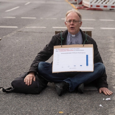 Blockade von "Scientists Rebellion" in München: Der Jesuiten-Pater Jörg Alt sitzt mit einem Schild mit der Aufschrift "Die Klimakatastrophe macht bis 2070 über 3 Milliarden Menschen zu Geflüchteten" um den Hals auf der Straße.