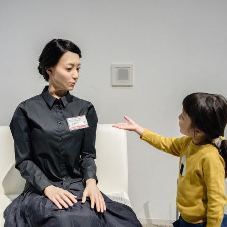 Ein junges Mädchen streckt einem weiblich aussehenden Roboter die Hand entgegen.
