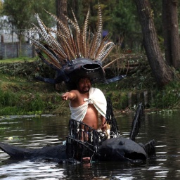 Ein Mann mit vielen Federn als Kopfschmuck sitzt in einem Boot in Form einer Amphibie: ein Axolotl in Mexiko