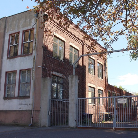 Bildbeschreibung: Das Eingangstor zu einem ehemaligen Betriebsgelände. Auf der linken Seite ist ein Verwaltungsgebäude zu sehen.