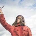 Ein Junge in Pilotenkostüm hält einen Papierflieger in die Luft.