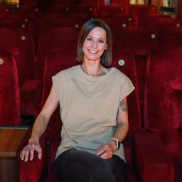 Sängerin Christina Stürmer trägt ein T-Shirt in der Farbe beige. Sie lächelt und sitzt in einem Kinosaal voller samtiger roter Sitze. An ihrem linken Arm sind Tattoos zu sehen.