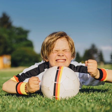 Junge im Trikot der deutschen Fußballmannschaft liegt im Gras, vor sich einen Ball, und drückt die Daumen: Den Ausdruck kannten offenbar schon die alten Römer, für die das Daumendrücken zu tun hatte mit &#034;jemandem Glück wünschen&#034;, &#034;jemanden in Gedanken unterstützen&#034;.