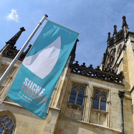 Ein Banner mit dem Motto "Suche Frieden" und dem Logo des Katholikentages hängt vor dem Dom in Münster.