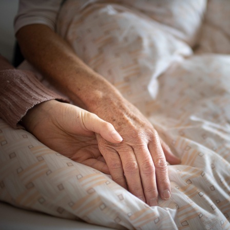 Eine ältere und eine jüngere Person halten sich an den Händen auf einer Bettdecke.