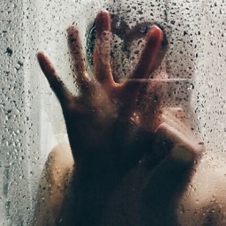 Eine Frau, die sich in einer Dusche befindet und mit den Fingern ein Herz an die Glaswand der Dusche zeichnet, die mit Wassertropfen besetzt ist.