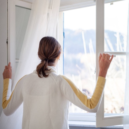 Eine Frau steht am Fenster und öffnet es weit mit Blick nach draußen: Richtiges Lüften hängt von den Umständen ab. Wie warm und feucht ist die Wohnung? Sind die Räume gut gedämmt? Trotzdem gibt es ein paar Grundregeln, die man beachten kann.