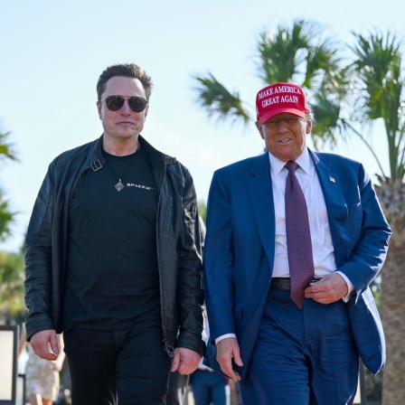Donald Trump und Elon Musk laufen unter Palmen nebeneinander her. 