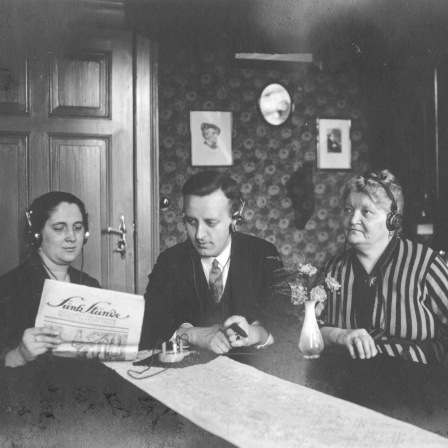Drei Radiohörer, zwei Frauen, in der Mitte ein Mann, mit Kopfhörern und Detektorempfänger. Undatiertes Foto aus den 1920ern. Die Frau links im Bild hält ein Papier, vermutlich ein Programmheft, mit dem Titel &#034;Funk Stunde&#034; in der Hand.