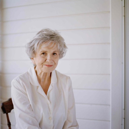 Alice Munro trägt eine weisse Bluse und sitzt auf einer Terasse vor einem weissen Holzhaus. Sie lächelt freundlich in die Kamera.