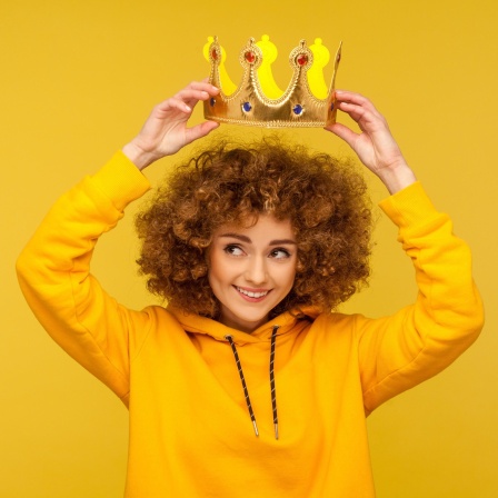 Eine junge Frau mit Locken setzt sich lächelnd eine Krone auf den Kopf