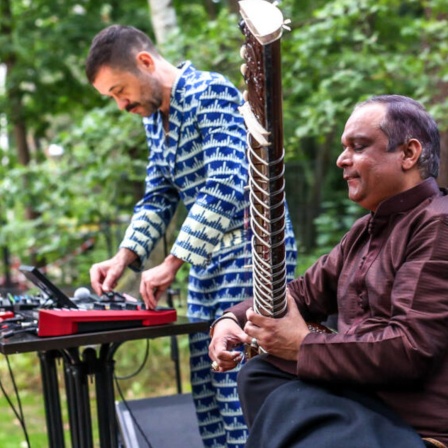 Zwei Musiker bei einem Open Air Auftritt. Einer spielt Sitar, der andere steht im Hintergrund an einem Mischpult.