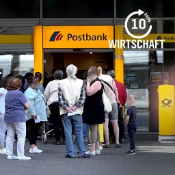 Kunden stehen in einer Warteschlange vor einer Filiale einer Postbank. 